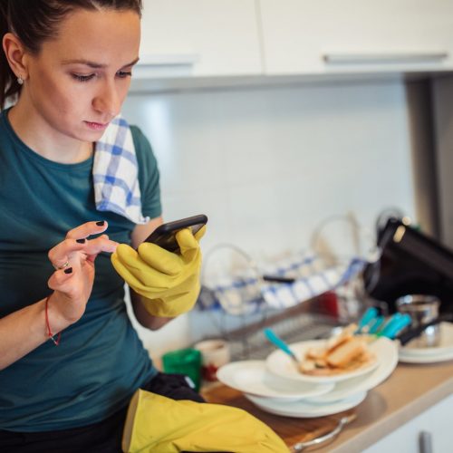 Young housewife is sitting on the kitchen counter, wearing protective gloves, and text messaging.